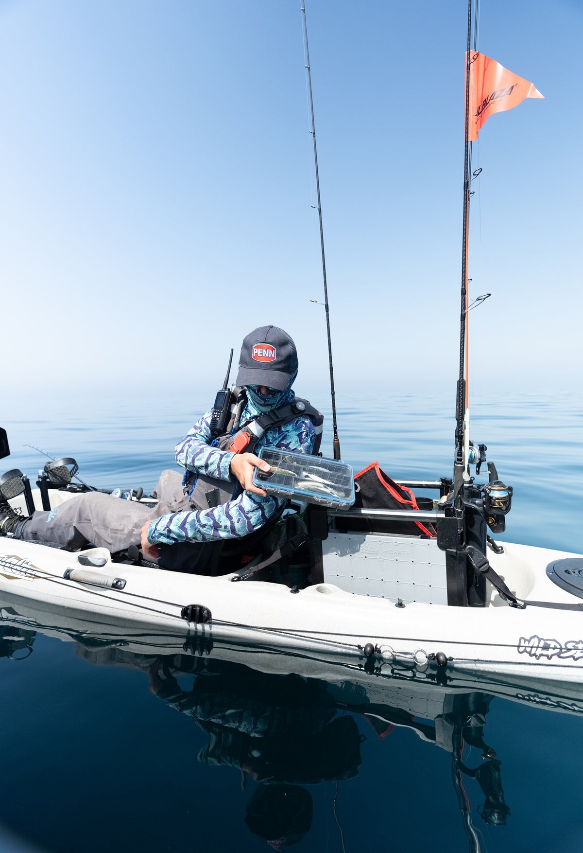 A person in a camouflage fishing jacket sits in a white sit-on-top fishing kayak on open water, putting a Plano StoAway Takle box back into his tackle box. The kayak has a mounted orange flag, fishing rods, and tackle gear, with a clear blue sky and calm water in the background.