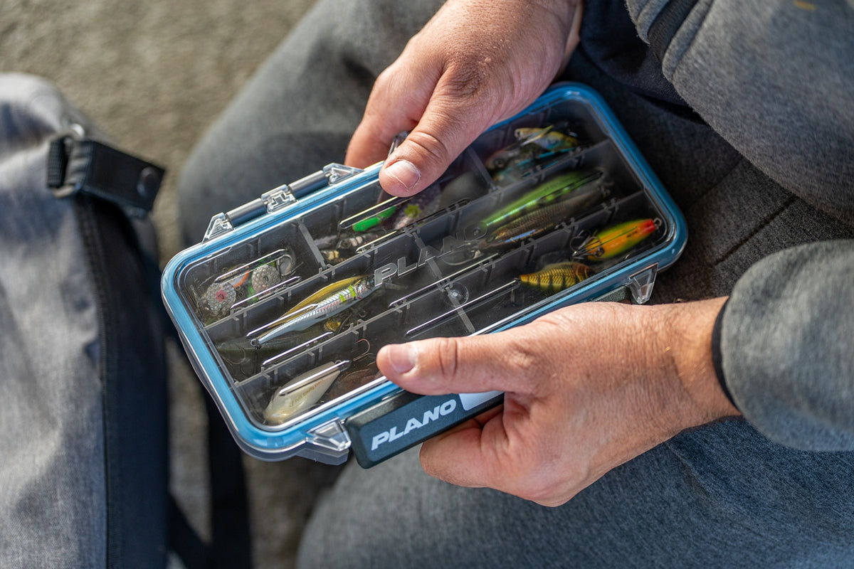 A close-up of a person holding an open blue and dark grey PLANO StowAway tackle box filled with fishing lures. Their hands holding the front, opening the latch with one hand, and legs visible against a grey background.
