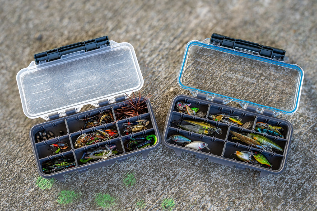 Two open Plano StowAway tackle boxes filled with fishing lures with divided compartments in various configurations to show the versatility. The left box has has a transparent lid, the right box has a transparent lid with blue outline showing the waterproofing. The boxes sit on a textured grey concrete