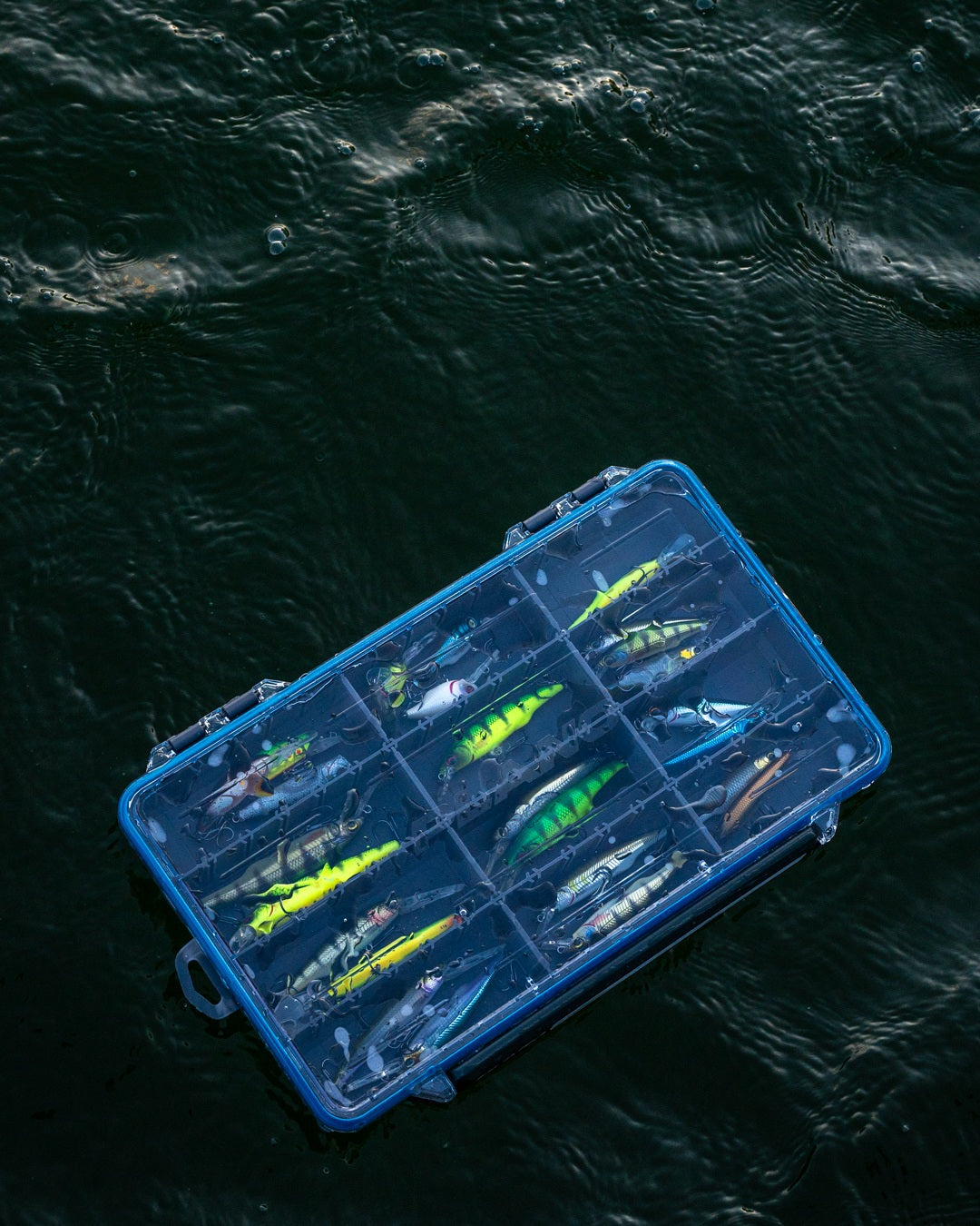 A Plano Waterproof StowAway tackle box filled with fishing lures, photographed from above floating on dark rippling water.