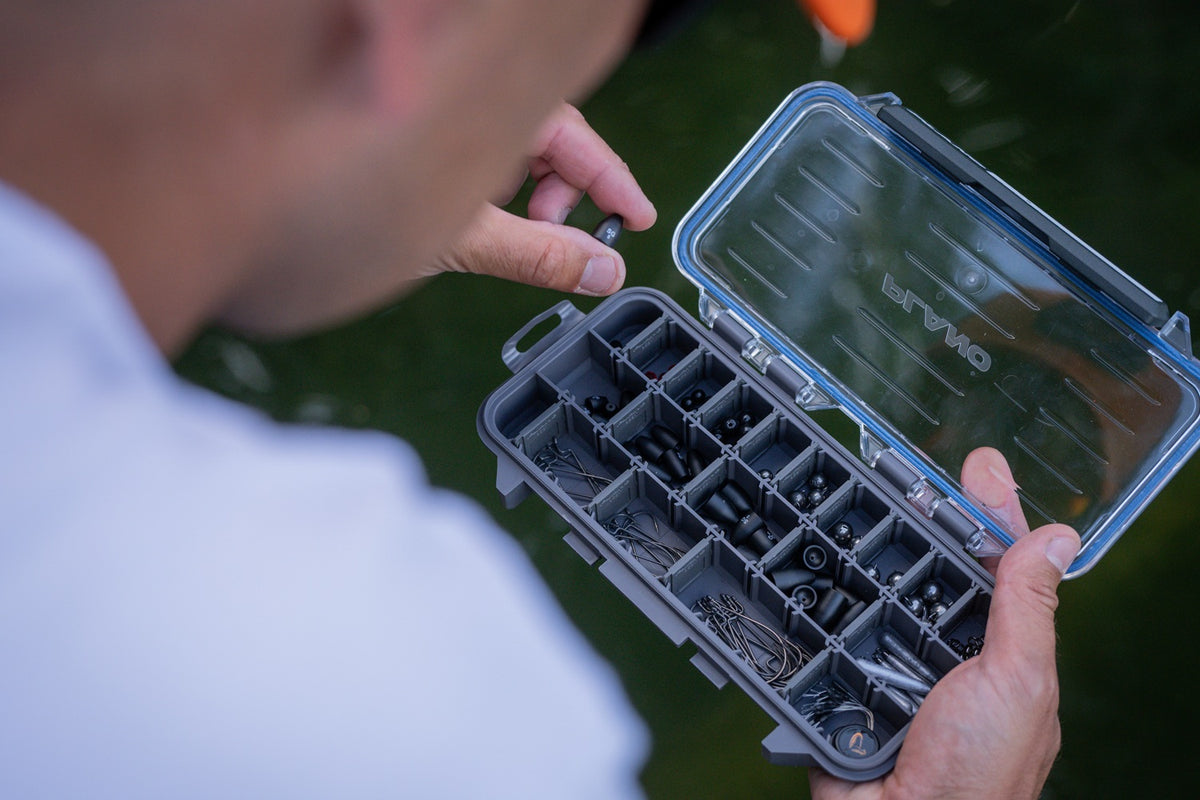 A close-up lifestyle shot of a person holding an open Plano StowAway Tackle Box filled with fishing hooks and bait, with a blurred outdoor background.