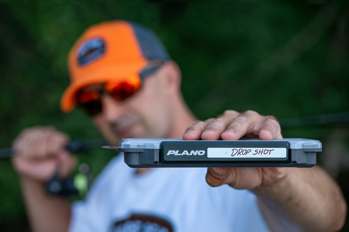 A blurred man in the background wearing an orange and gray cap holds a Plano StowAway side on displaying the latch area with Drop Shot written into the labelling area in the foreground, with a further distanced blurred outdoor background of green foliage.